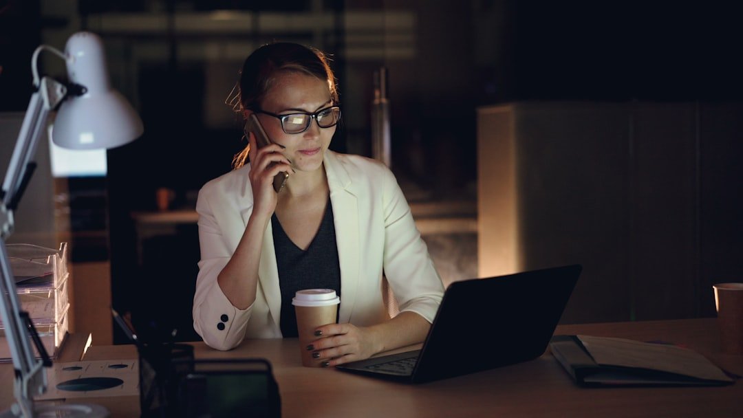 Work messages after hours: Woman talking on phone at desk with laptop and coffee