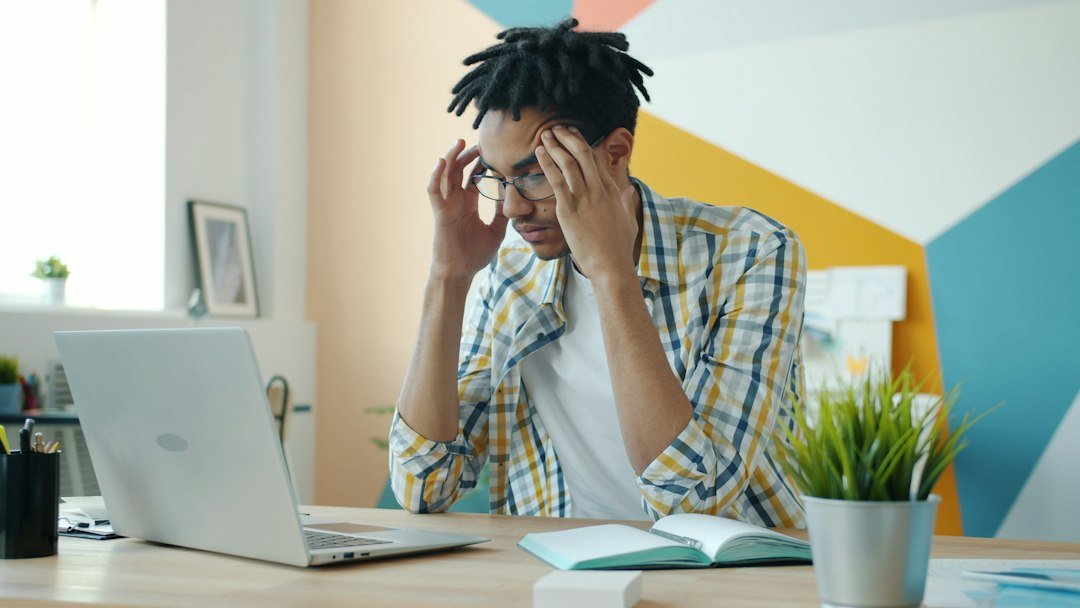 Man with dreadlocks holding head at desk with laptop